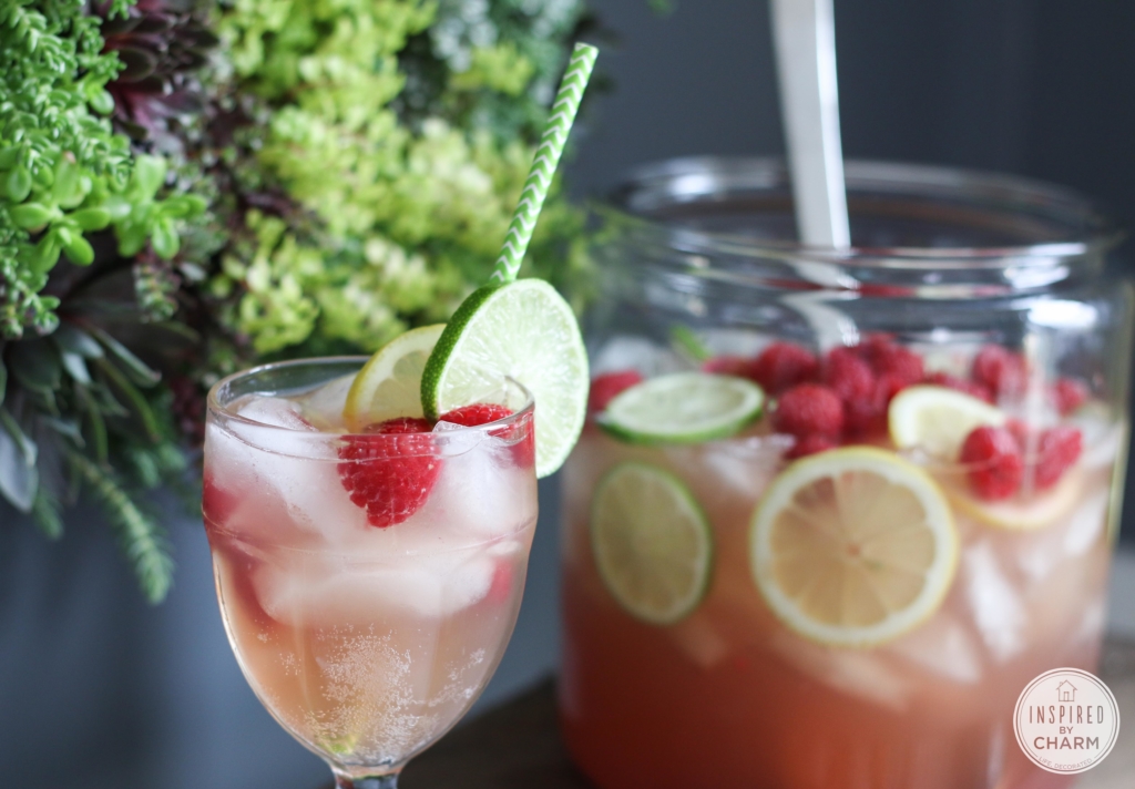 Raspberry Berry Cocktail in a stemmed glass with paper straw and citrus slices and large pitcher in the background.