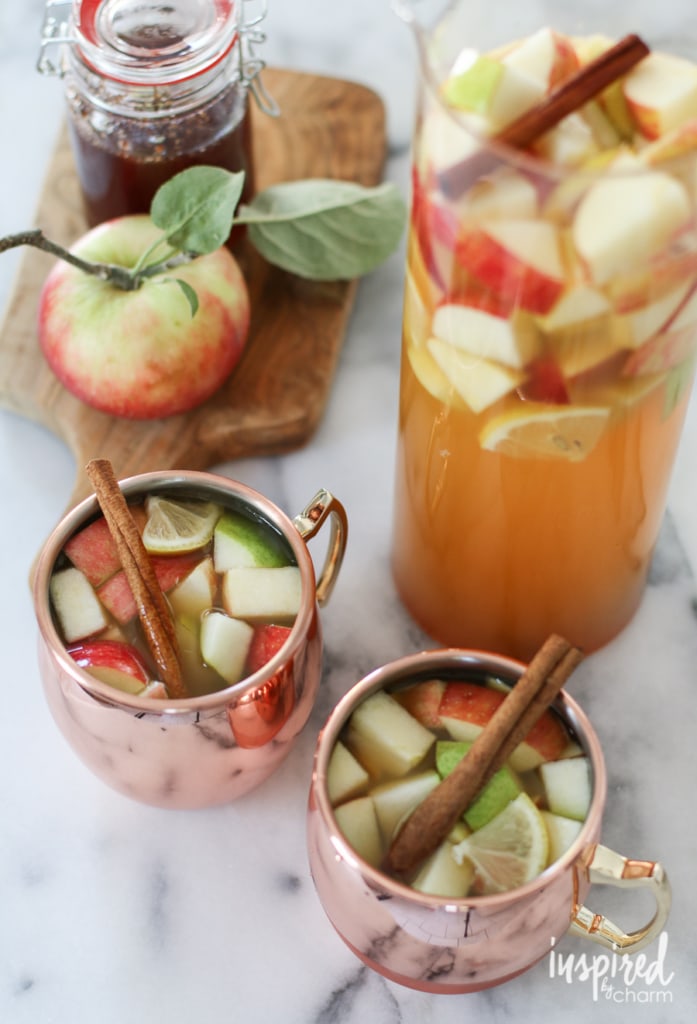 two copper mugs filled with apple cider sangria next to a pitcher and an apple.