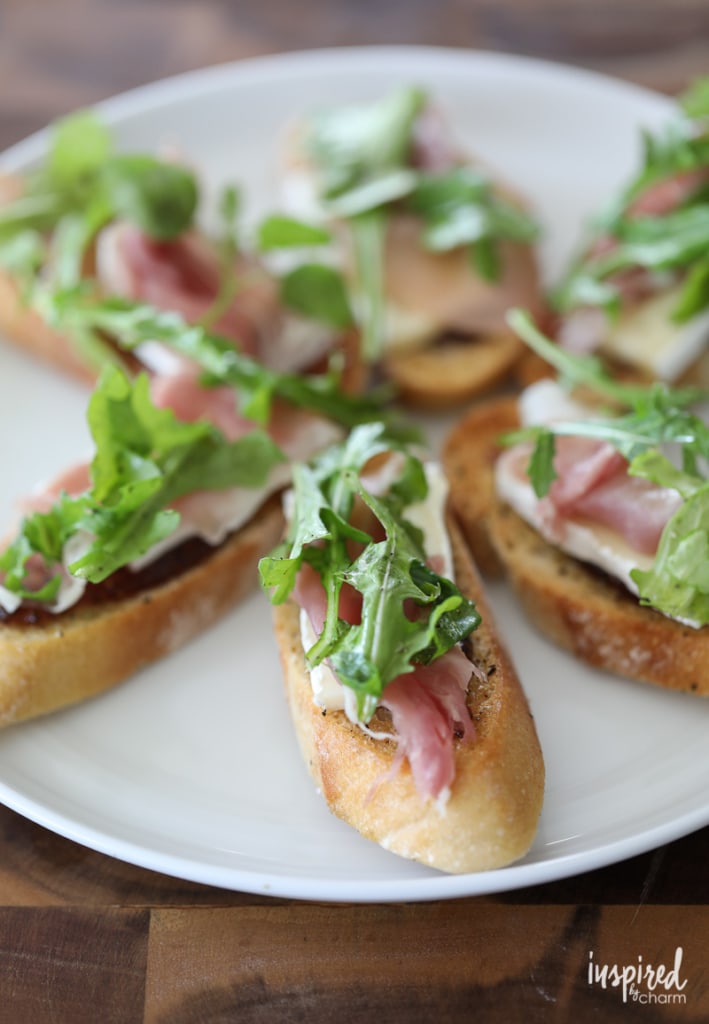 Plate of crostinis topped with brie, fig, prosciutto, and arugula lettuce.