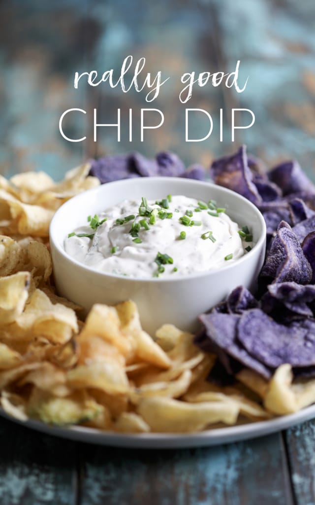 Chip dip in a white bowl surrounded by potato chips on one side and blue corn chips on the other