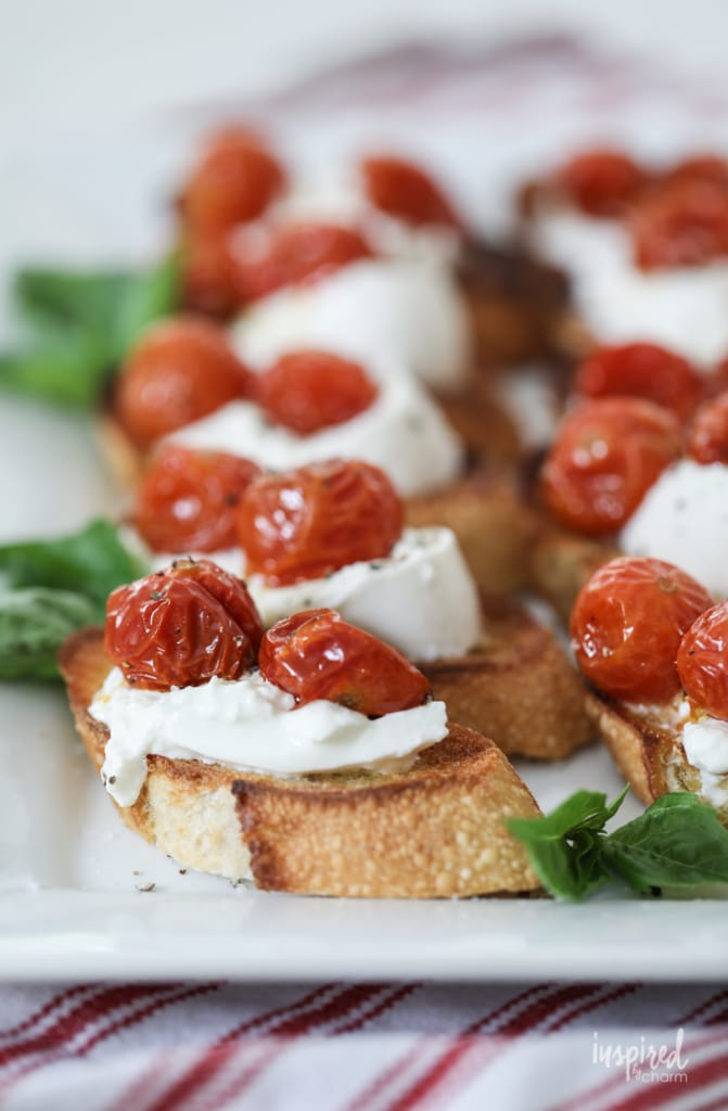 Roasted Tomato and Burrata Crostini on a white platter with basil leaves.