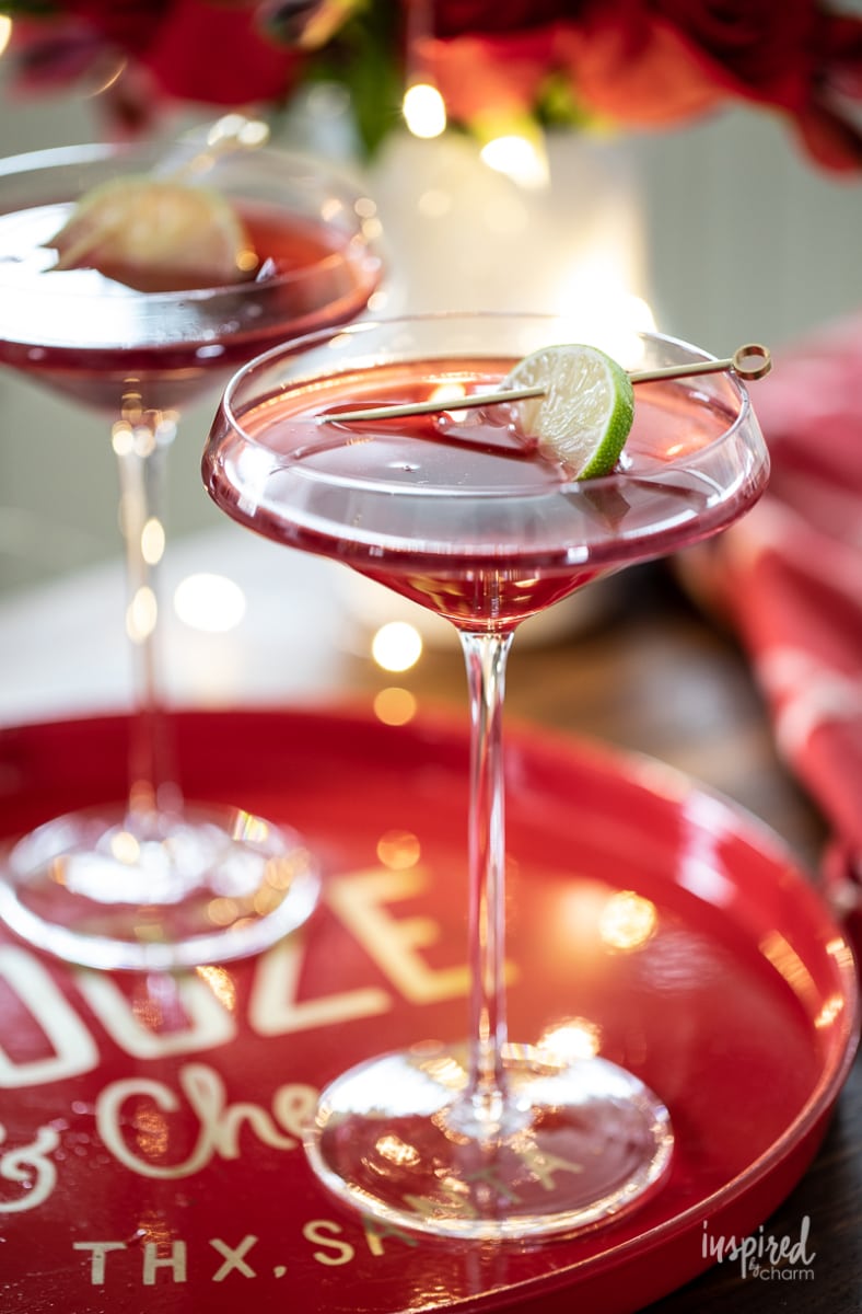 very tall stemmed martini glass with red Christmas cocktail and lime garnish 