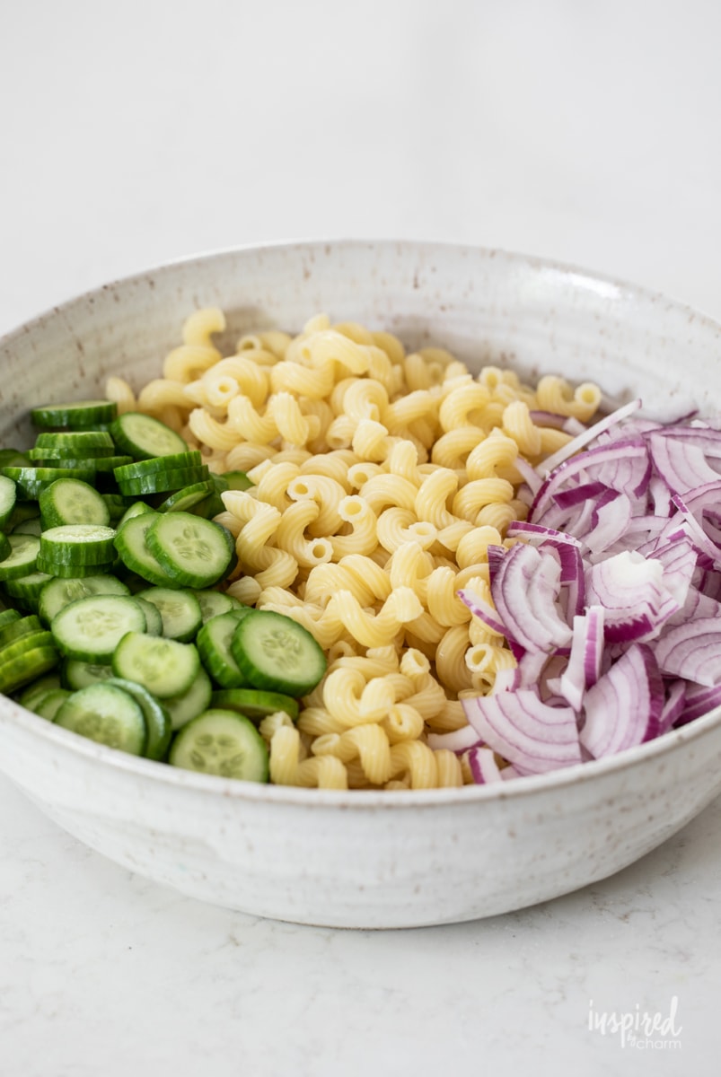 a large bowl with sliced cucumbers, cooked pasta, and red onions.
