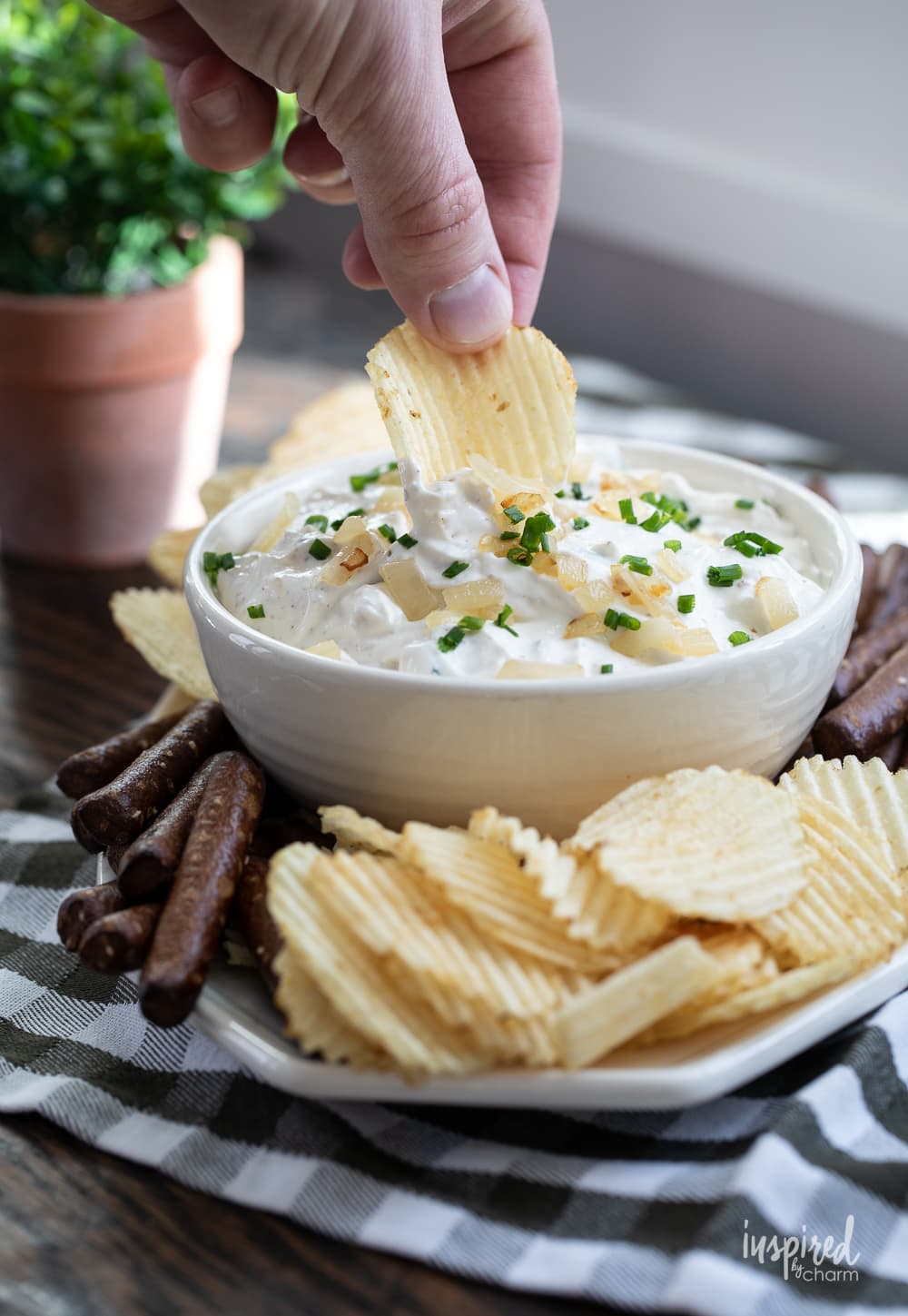 hand dipping into sour cream and onion dip in a bowl with potato chips and pretzels.