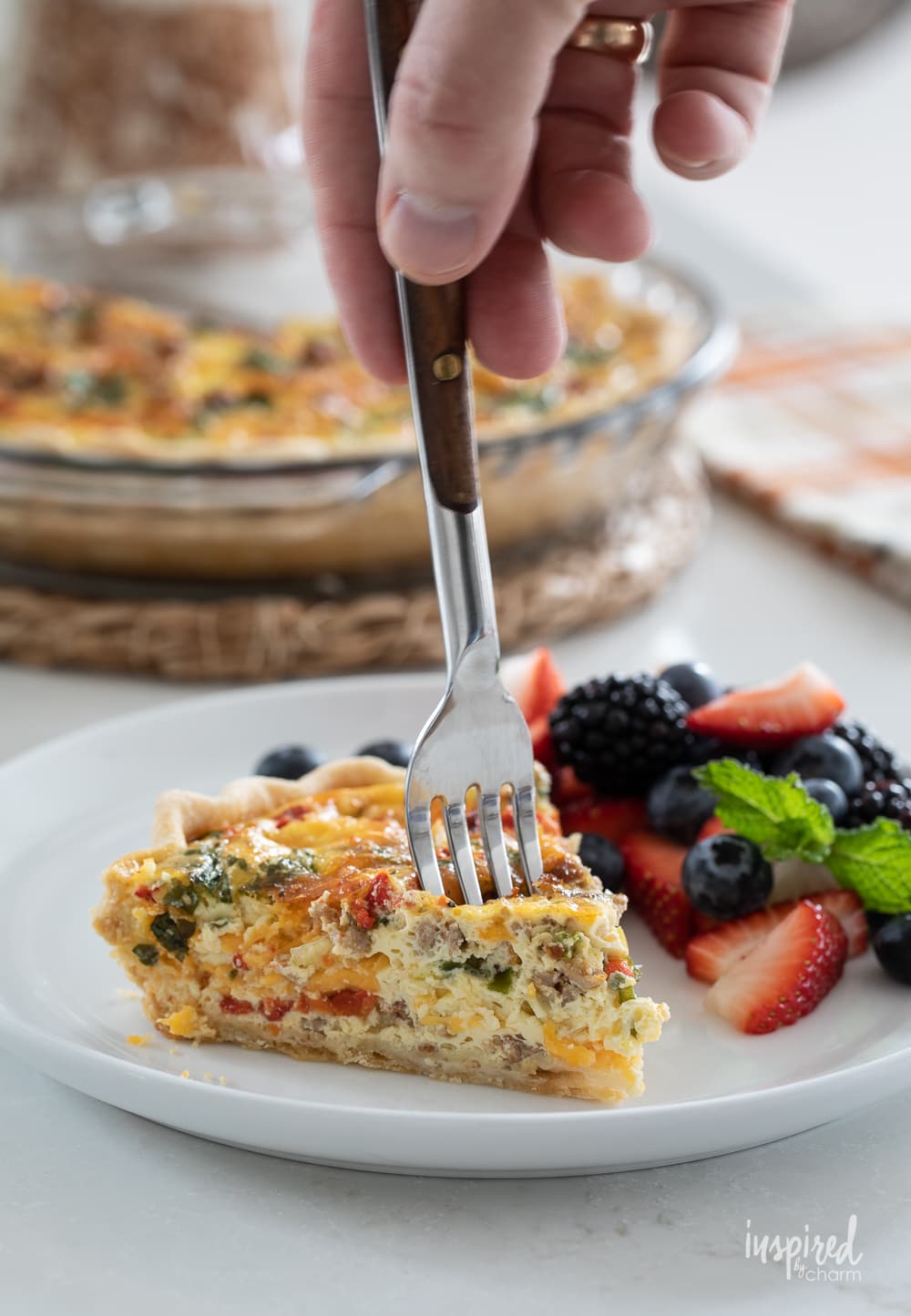 hand holding a fork going into a slice of red pepper and sausage quiche on a plate with fresh berries. 