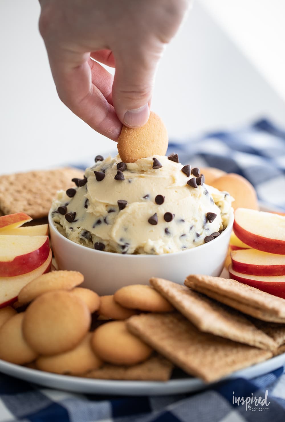 hand dipping Nilla wafer into chocolate chip cheesecake dip in a bowl with cookies and apple slices.