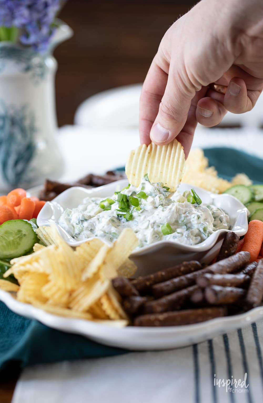 hand with chip in green onion dip in a bowl on a plate with chips and vegetables.
