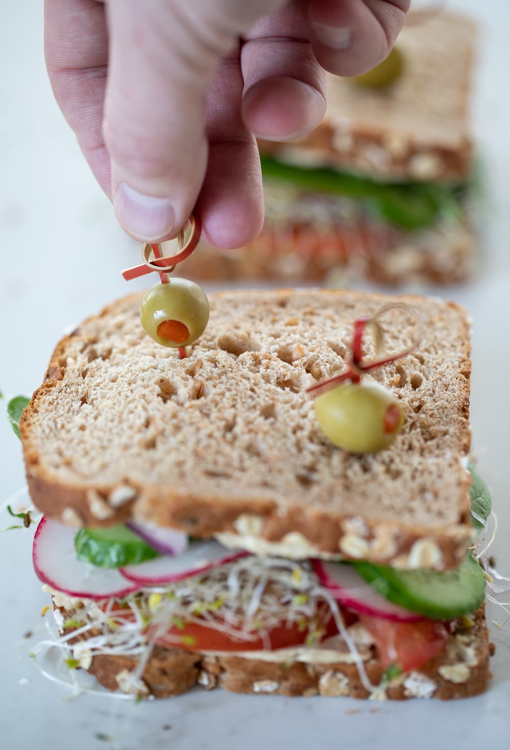 hand putting olive and toothpick into hummus sandwich.