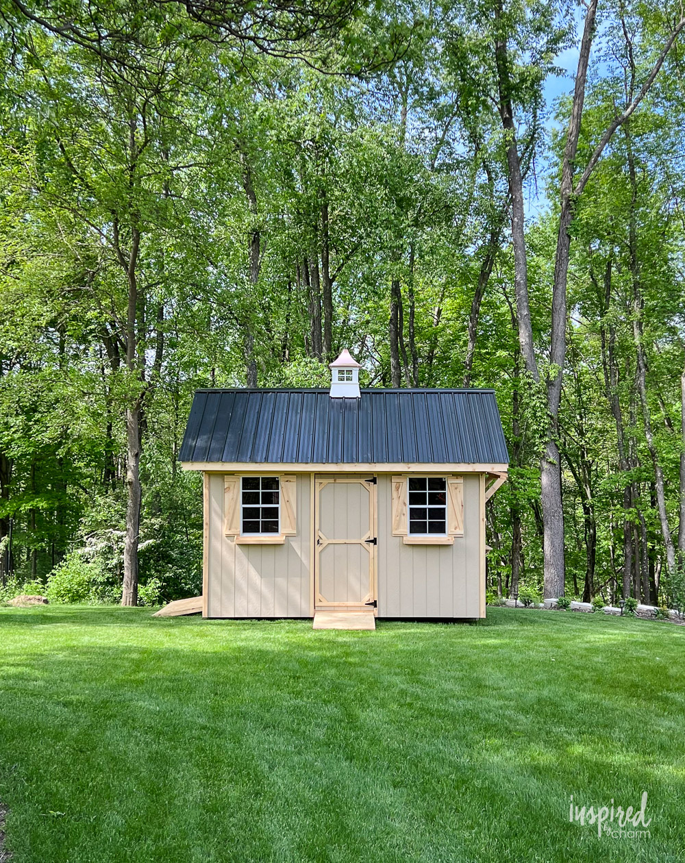 wood shed on top of green grass with trees behind it.