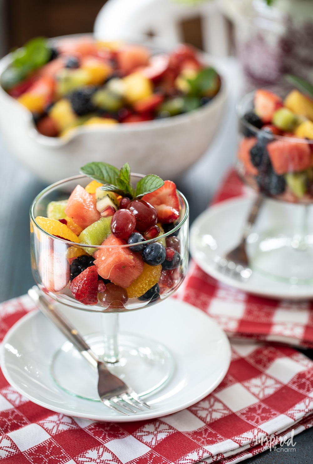 fruit salad in a glass footed dish on a plate with a big bowl of fruit.