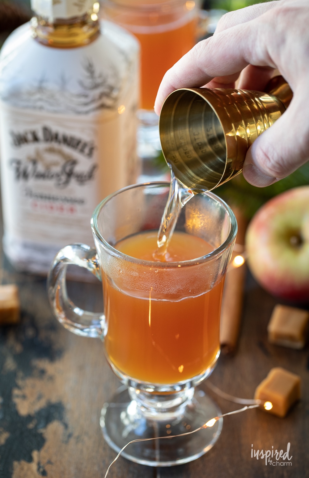 hand holding a jigger adding whiskey to glass mug of apple cider.