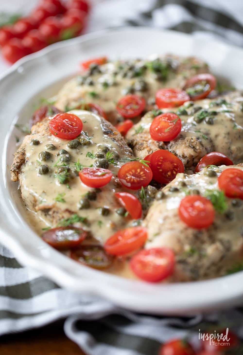 a platter filled with chicken breasts covered in a caper cream sauce and garnished with cherry tomatoes.