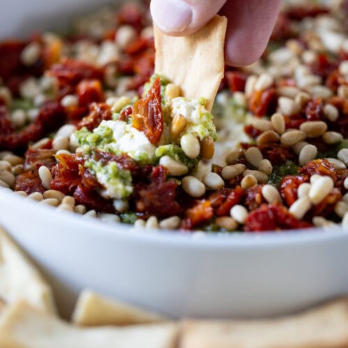 hand with pita chip dipping into Pesto and Feta Layer Dip.