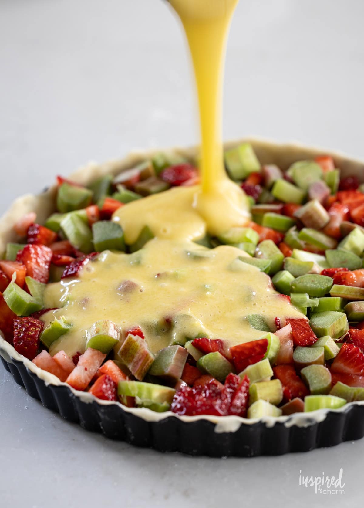 custard being board over strawberries and rhubarb in tart pan.